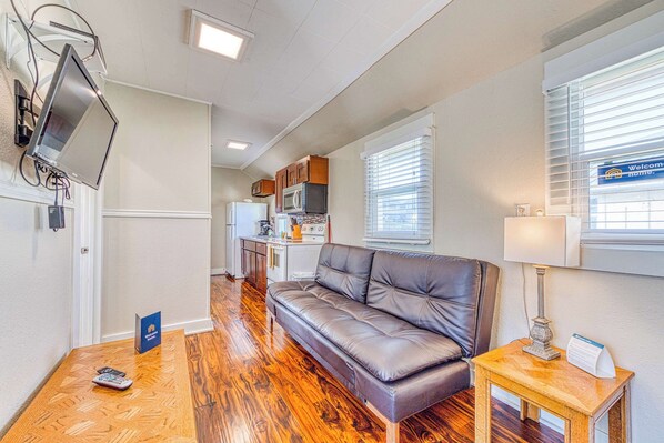 A compact living area with a sofa, a small table, a TV mounted on the wall, and a kitchen in the background.