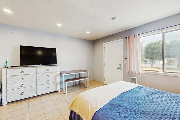 Un dormitorio con una cama, televisor, cómoda, ventana con cortinas y una puerta.