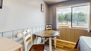 Una stanza con un tavolo da pranzo in legno, sedie bianche, una finestra con tende e un letto con copriletto decorato.