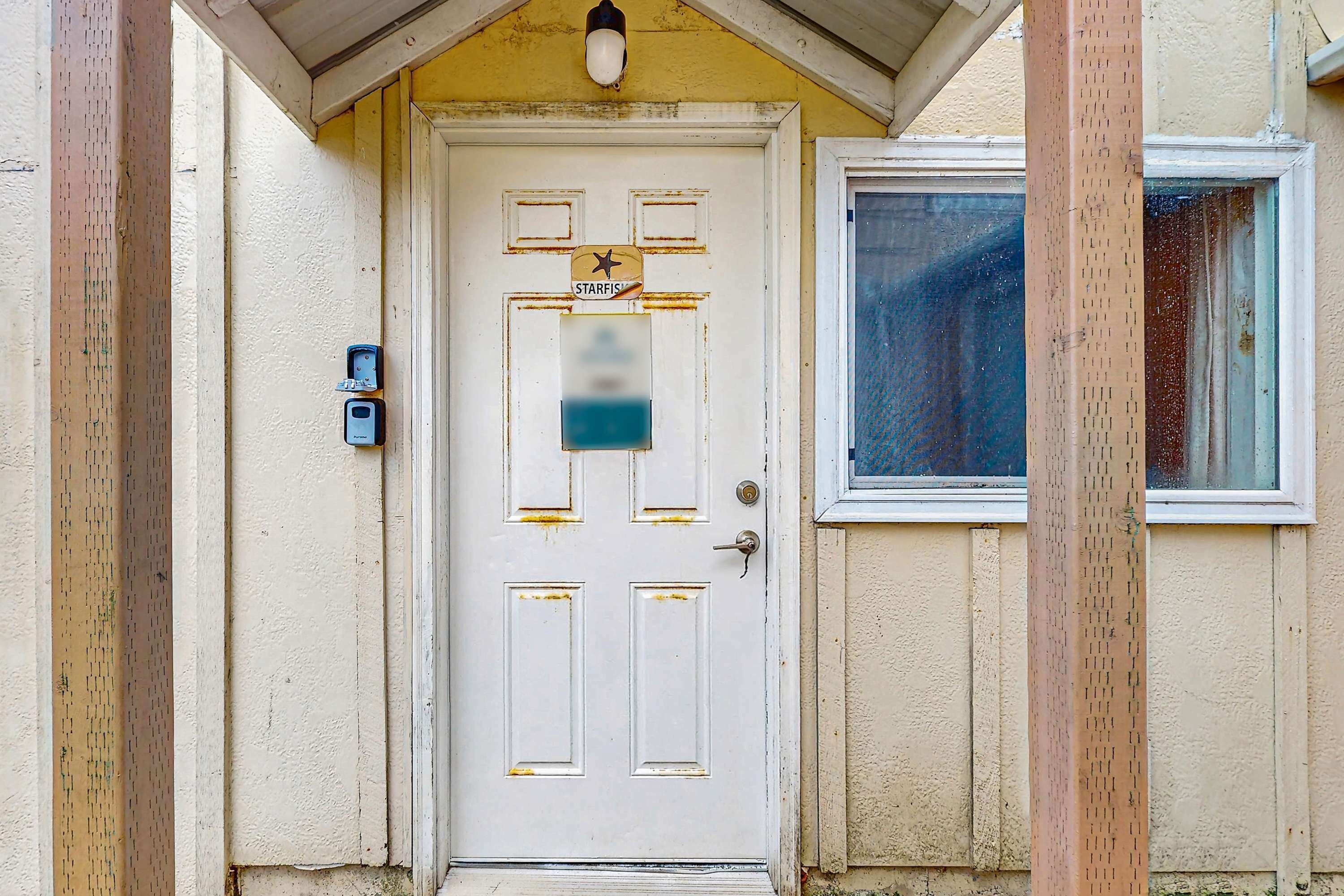 Eine weiße Tür mit einem Schild in Form eines Seesterns und einem blauen Aufkleber, ein Fenster mit Insektenschutzgitter und ein Holzpfosten.