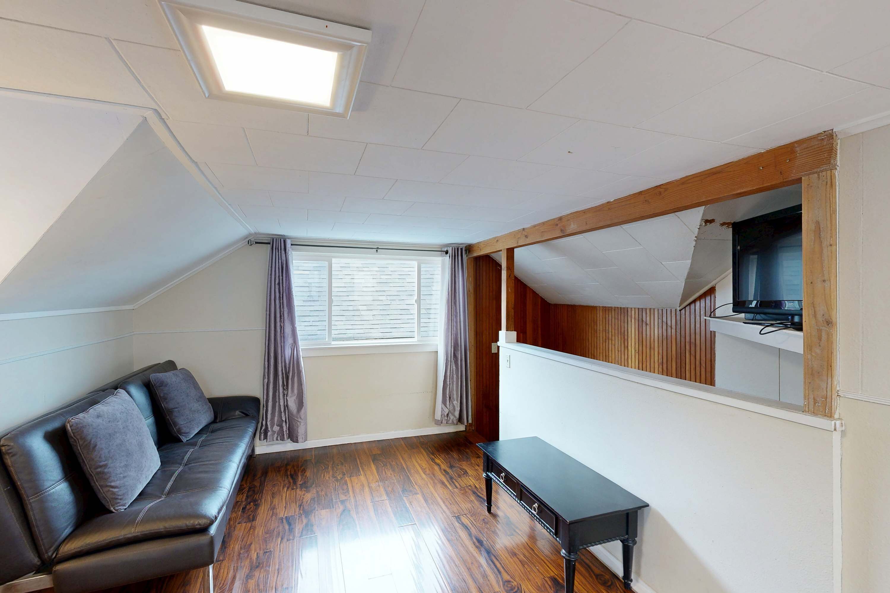Een kleine, goed verlichte kamer met een zwarte leren bank, een houten bankje en een televisie bevestigd aan een houten balk.