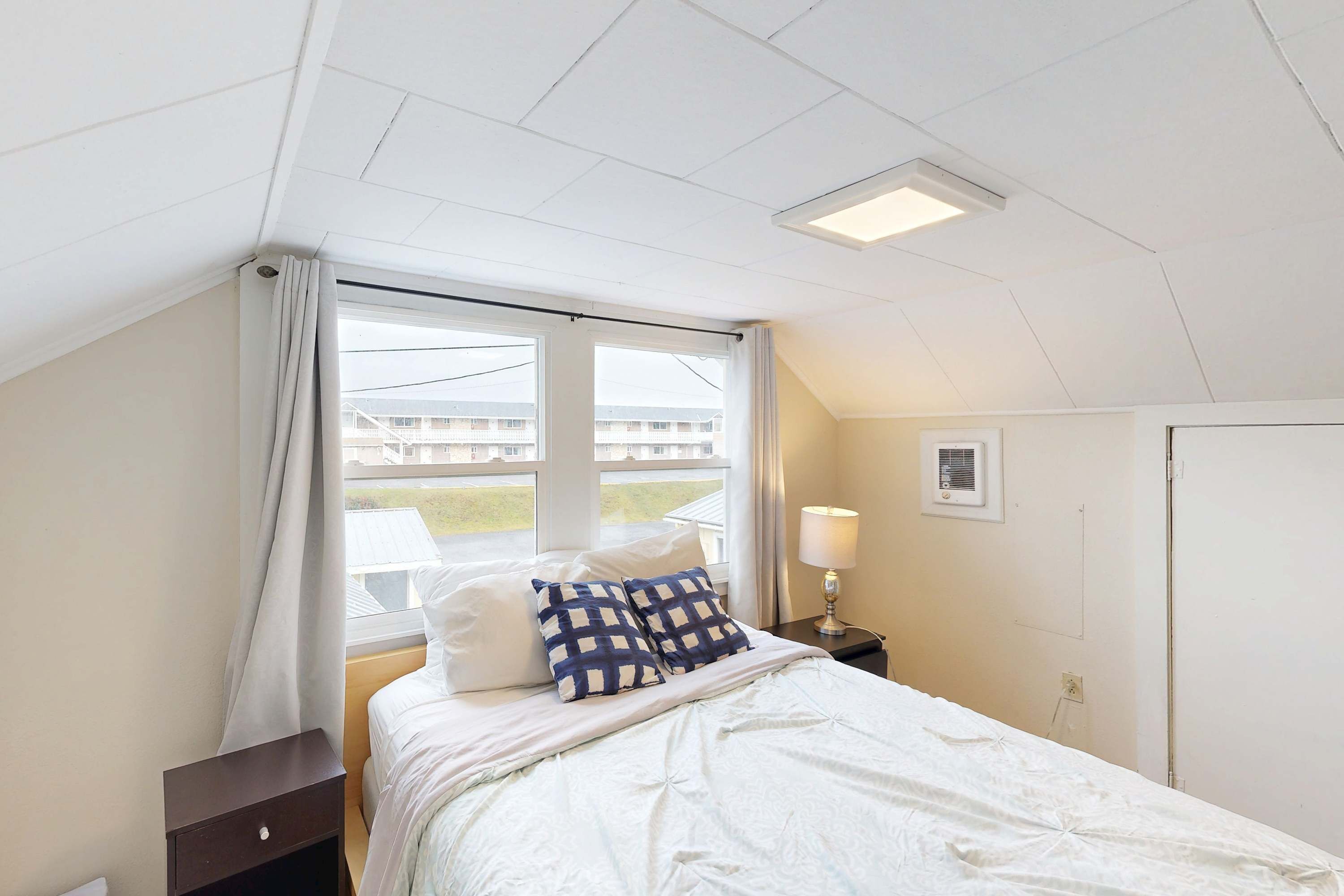 A bedroom with a bed, a nightstand, a lamp, a window with curtains, and a door.