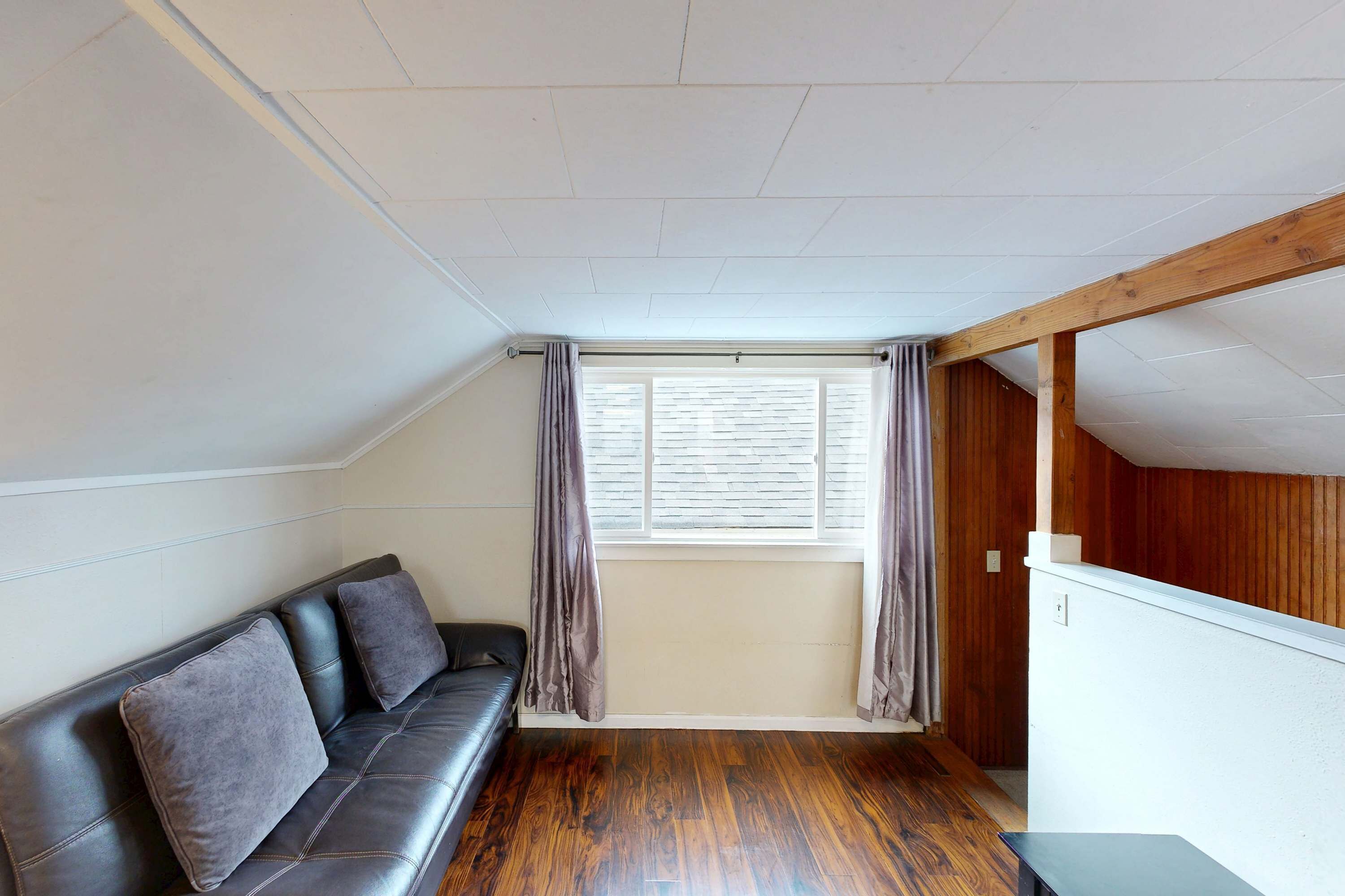 A room with a black leather sofa, a wooden floor, a window with curtains, and a sloped ceiling.