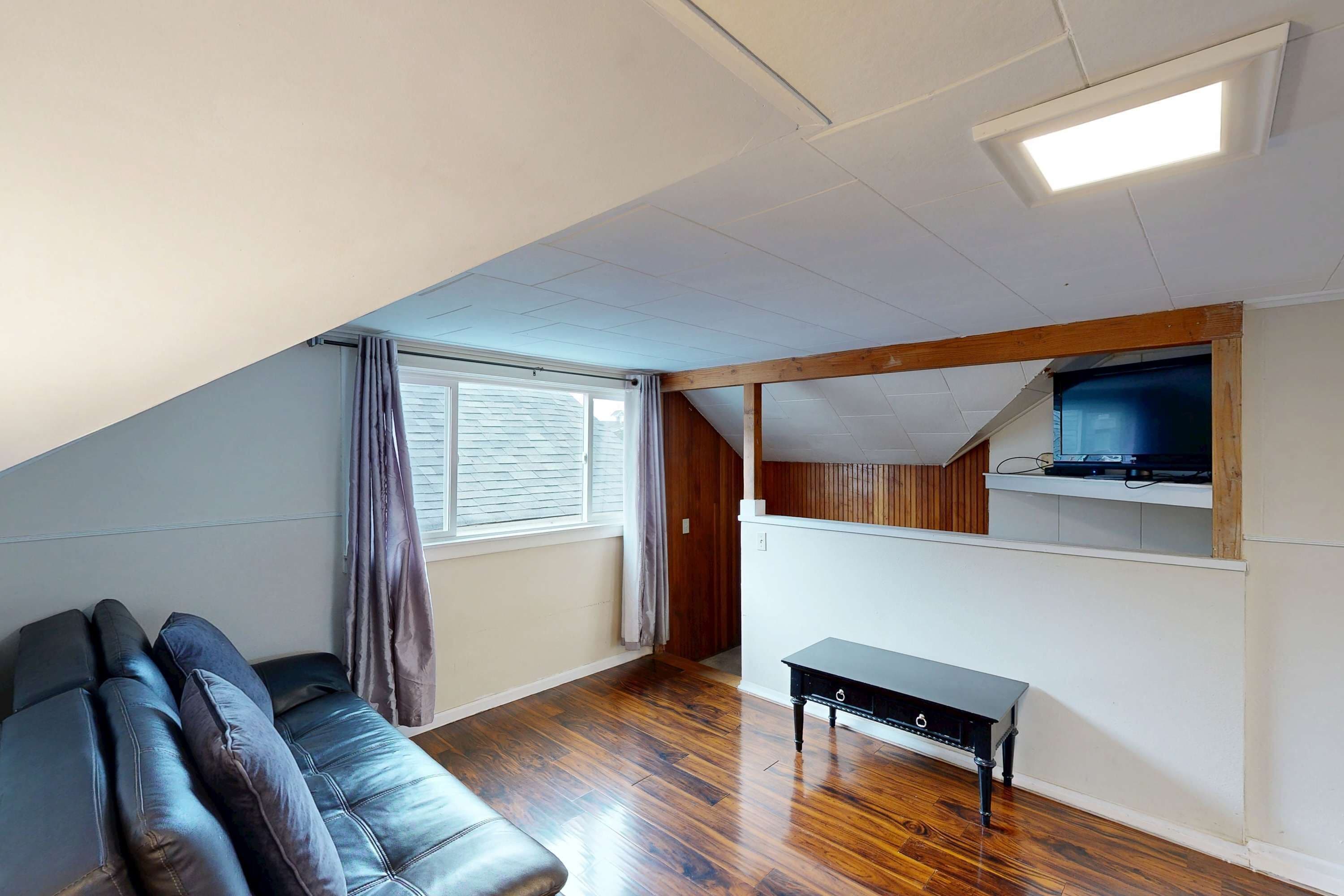 Una habitación con techo inclinado, un banco de madera, una mesa pequeña, un ventanal con cortinas y un televisor montado.