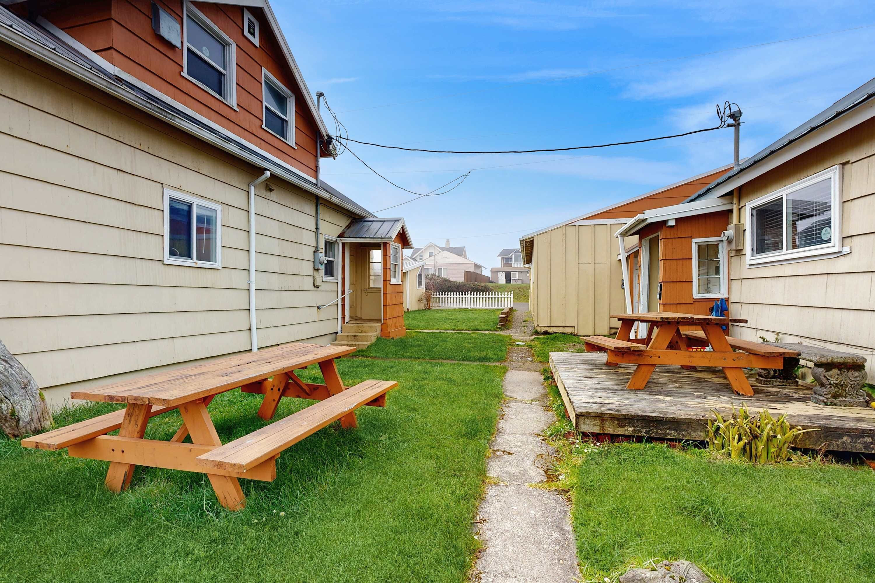 A residential area with two wooden picnic tables on a grassy lawn, a pathway, and a white picket fence in the background.