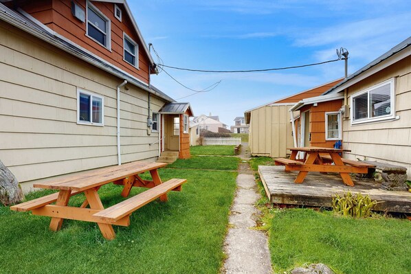 A residential area with two wooden picnic tables on a grassy lawn, a pathway, and a white picket fence in the background. - Conveniently Located Home with Access to Beach and Town One Dog Welcome (Rockaway Beach)