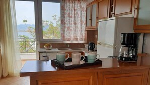 A kitchen with wooden cabinets, a refrigerator, a coffee maker, and a window with a view of trees and mountains. - Apartment 'A View From The Attic Window' with Sea View, Wi-Fi and Air Conditioning (Kyparissia)