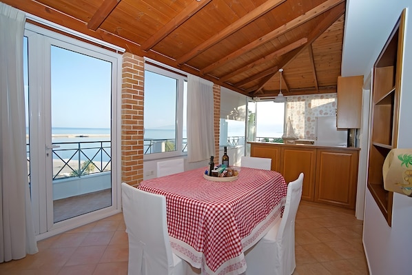 A dining area with a table set for two, a kitchen, and a balcony with a view of the sea.