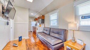 A compact living area with a sofa, a small table, a TV mounted on the wall, and a kitchen in the background. - Dog Friendly Triplex 3 private apartments Easy Walk to the Beach and Town (Rockaway Beach)
