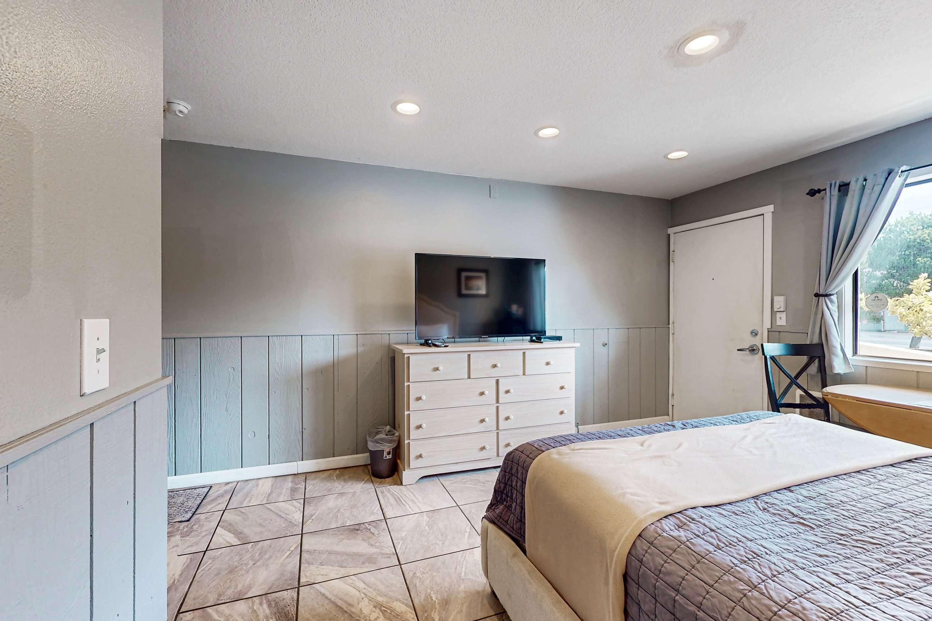A bedroom with a bed, a dresser, a television on top, a window with curtains, and a door.