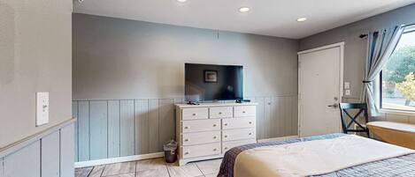 A bedroom with a bed, a dresser, a television on top, a window with curtains, and a door.