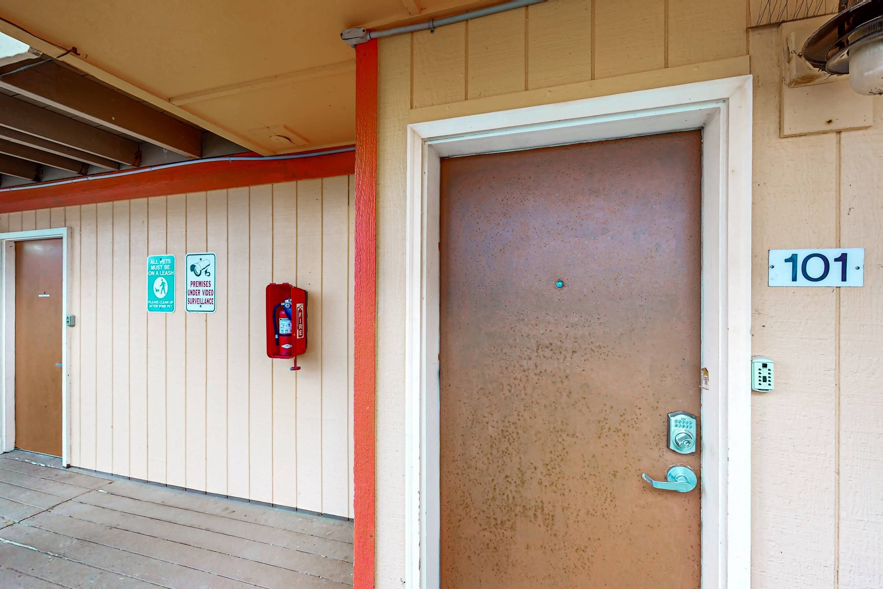 A building entrance with a closed door marked '101', a fire alarm, and signs indicating surveillance and a leash policy.