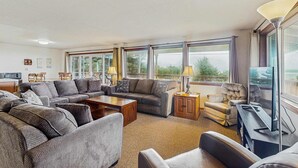 Ein geräumiges Wohnzimmer mit einer großen Eckcouch, einem Couchtisch und einem Fernseher. Mehrere Fenster sorgen für natürliches Licht und Blick ins Freie.