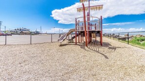 Uma área de recreação infantil com escorrega e estrutura de escalada, cercada por uma grade e uma área com brita.