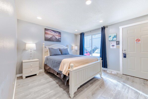 A bedroom with a bed, bedside tables, a window with curtains, a door, and a wall with a painting and signs. - Coastal room near the beach and downtown dog friendly (Rockaway Beach)