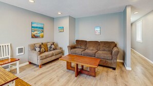 A living room with a beige sofa, a wooden coffee table, and a white chair. There are three wall-mounted paintings and a window with blinds. - Dog Friendly Room with Kitchen Located Near the Beach and Downtown (Rockaway Beach)