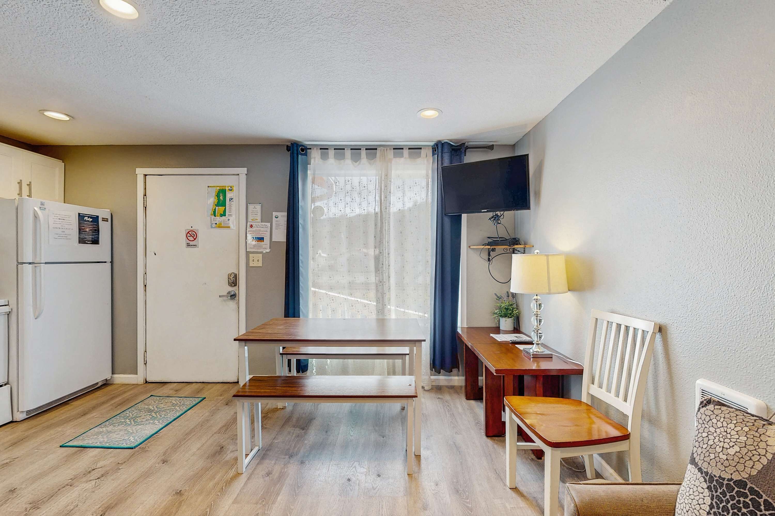 A compact kitchen with a refrigerator, a dining table with benches, a small table with a lamp, and a chair.