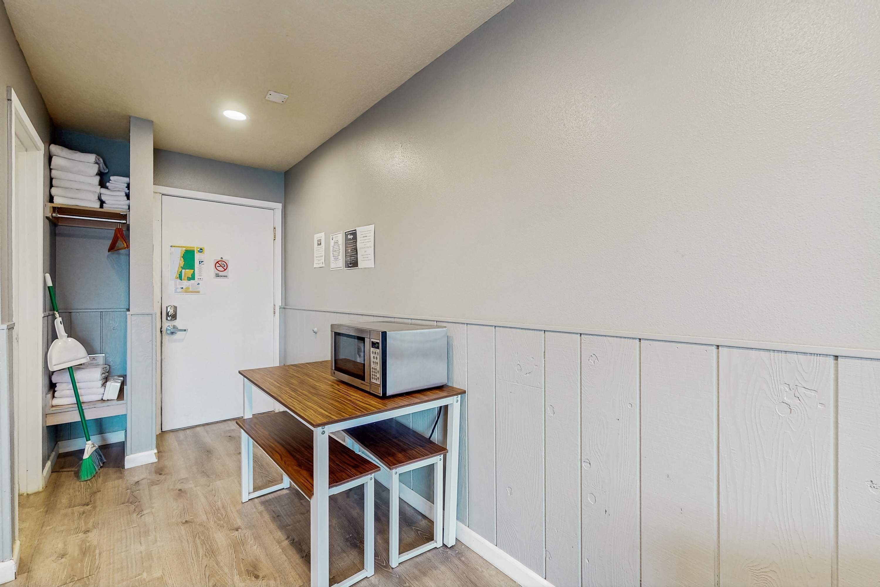 A small kitchenette with a microwave on a wooden table, a broom, and a shelf with towels.