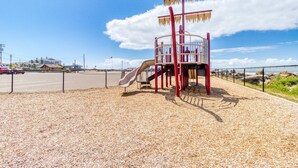 Un parque infantil con tobogán y estructuras para escalar, rodeado por una valla y un área de estacionamiento.