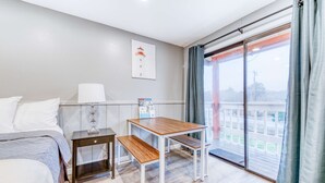 A hotel room with a bed, a small wooden table with benches, a nightstand, a wall-mounted lamp, and a large window with curtains. - Dog Friendly Room with Kitchen Located Near the Beach and Downtown (Rockaway Beach)