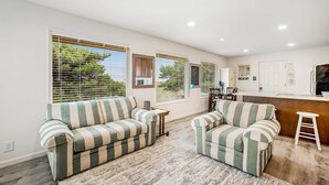 Ein geräumiges Wohnzimmer mit gestreiften Sofas, Holzboden und großen Fenstern mit Jalousien.
