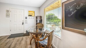 Una habitación con una mesa de vidrio, sillas de mimbre, una pizarra con la inscripción 'Welcome to Paradise North' y una puerta con ventanal, además de un tablón de anuncios.