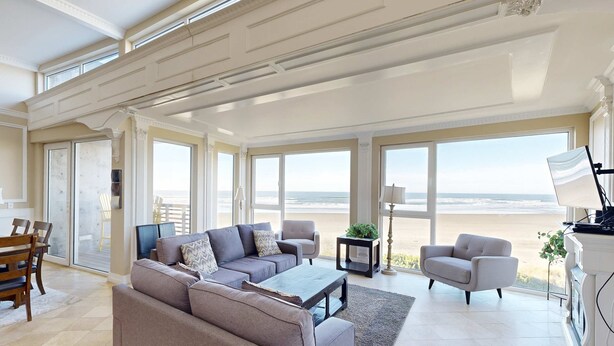 Een ruime woonkamer met een grote bank, fauteuils en een salontafel. Er zijn grote ramen met uitzicht op het strand.