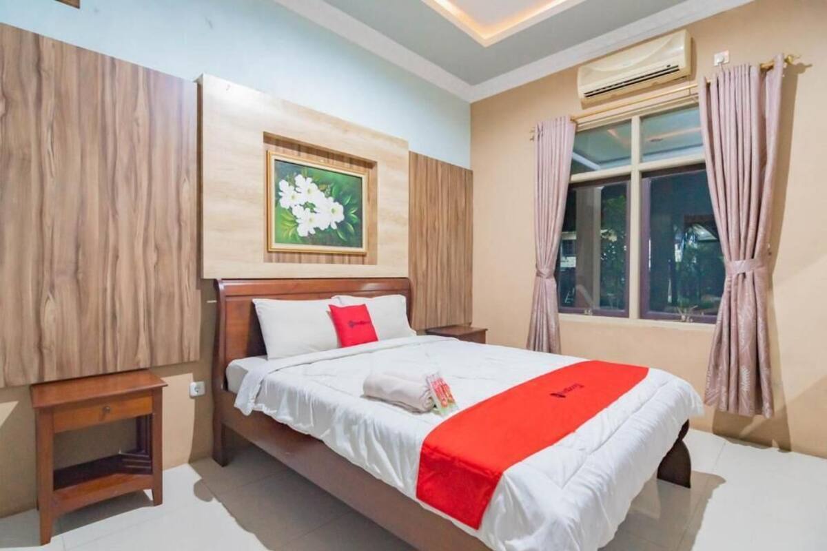 A hotel room with a wooden bed, a nightstand, a window with curtains, and a wall-mounted air conditioner.