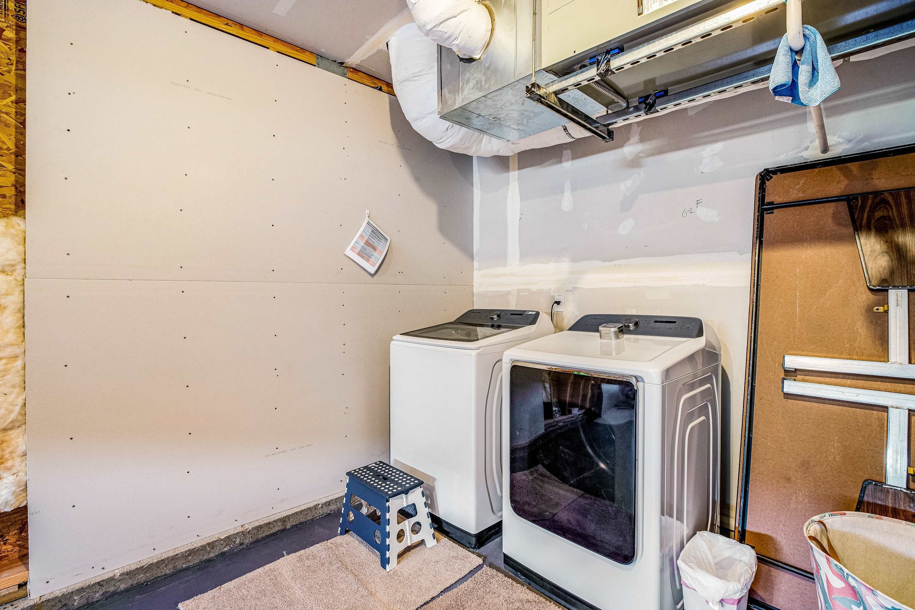 A laundry room with two stacked washers and dryers, a small stool, and a trash bin.