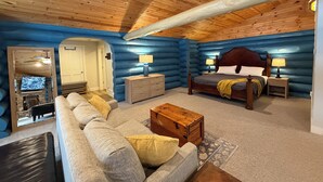 L’intérieur d’une cabane en rondins, avec un plafond en bois, un grand lit doté de deux tables de chevet, un canapé et un téléviseur placé dans un coin.