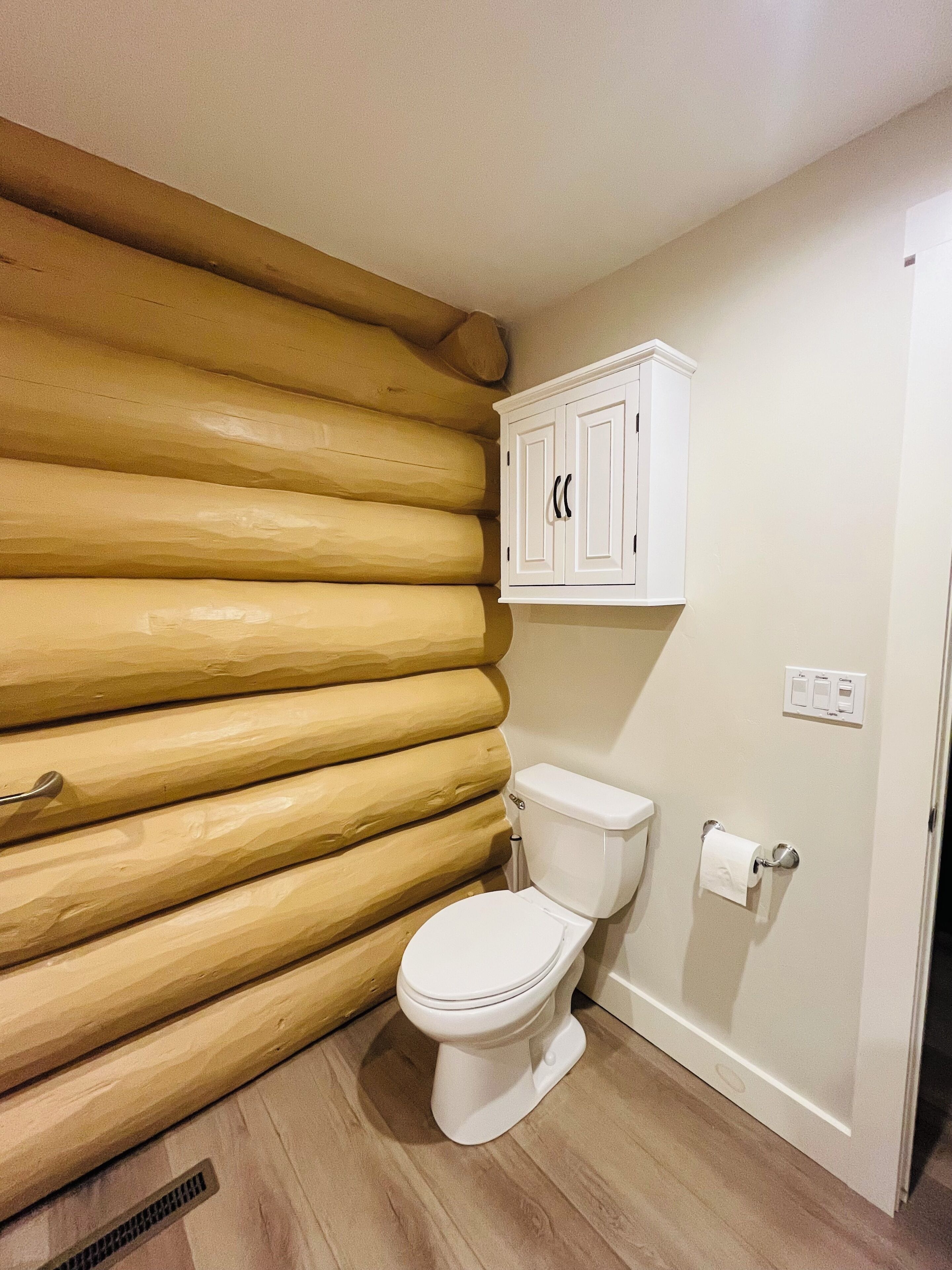 A bathroom with a toilet, wooden log wall, and a white cabinet above the toilet.