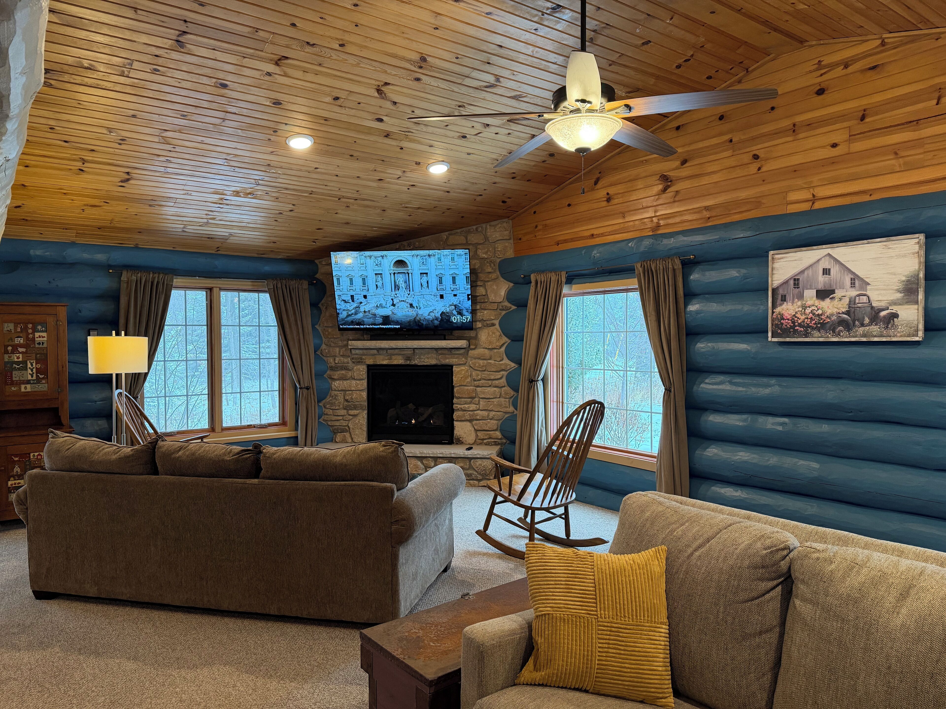A cozy log cabin interior with a fireplace, two sofas, a rocking chair, and a television mounted on the wall.