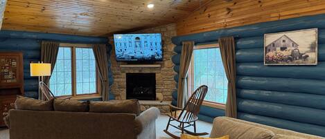 A cozy log cabin interior with a fireplace, two sofas, a rocking chair, and a television mounted on the wall.