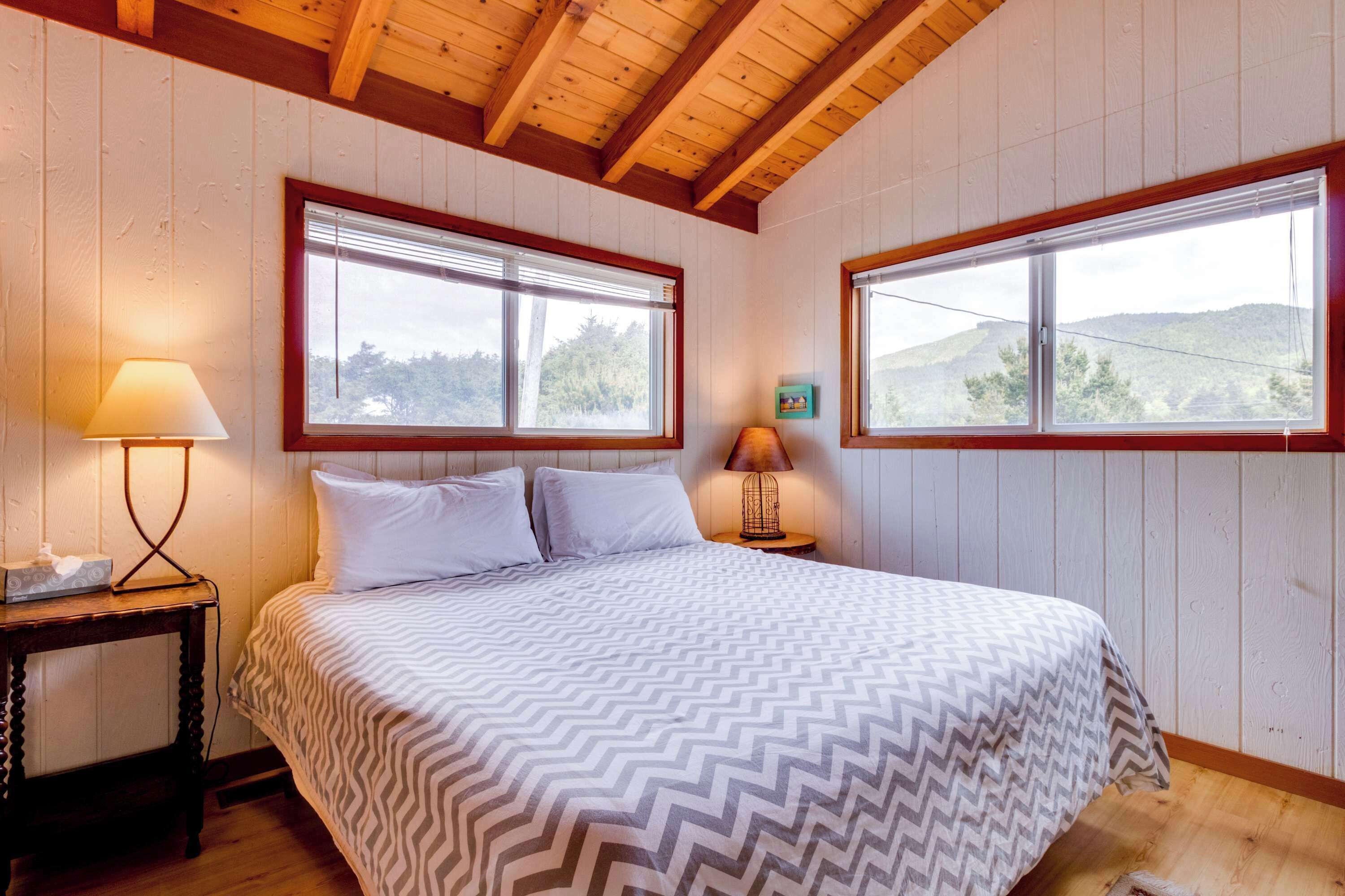 A bedroom with a bed, two windows, a nightstand, and a lamp.