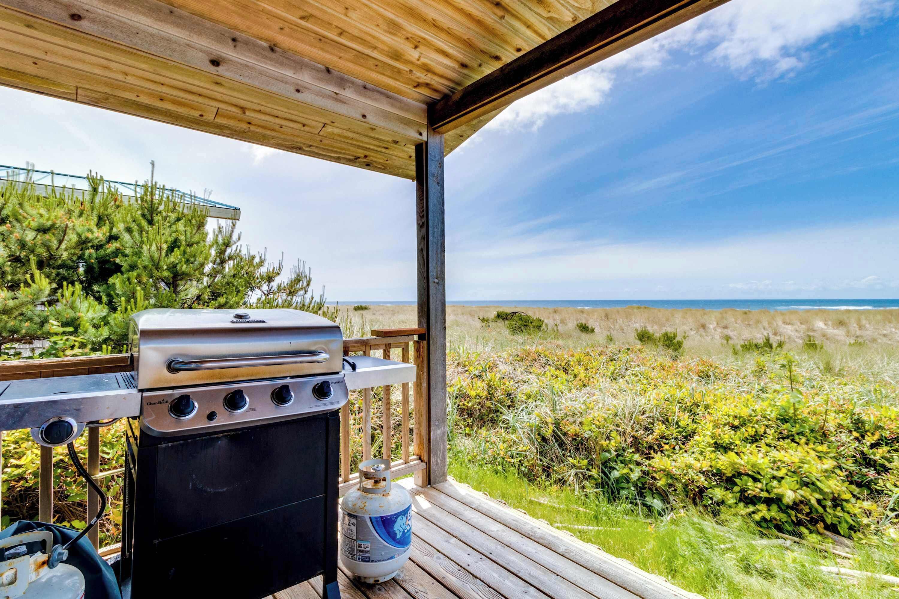 Ein überdachter Außenbereich mit Grill und Propangasflasche, mit Blick auf Strand und Dünen.
