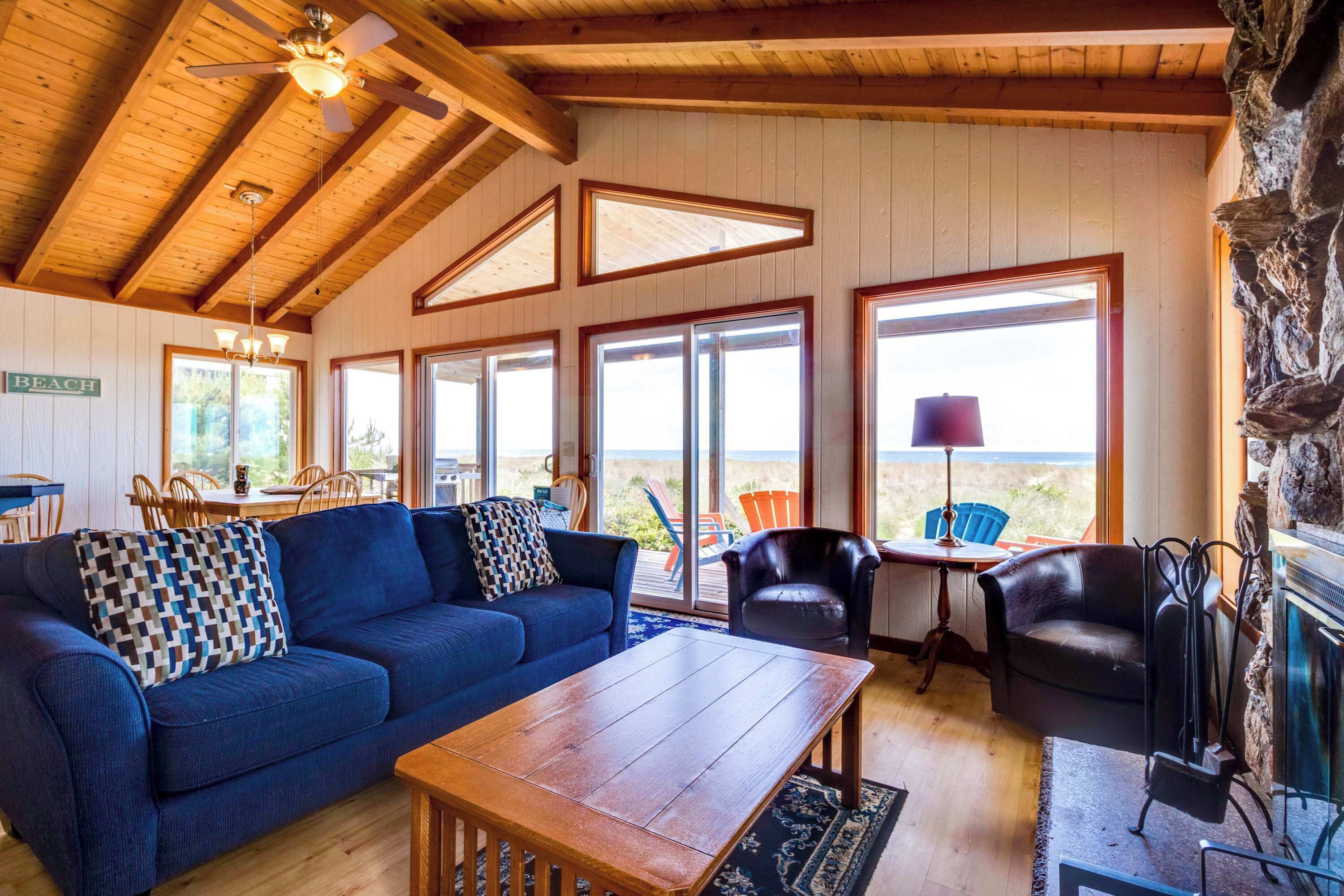 A cozy living room with a blue sofa, wooden coffee table, and a stone fireplace. Large windows offer a view of the beach.