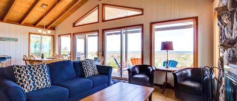 A cozy living room with a blue sofa, wooden coffee table, and a stone fireplace. Large windows offer a view of the beach.