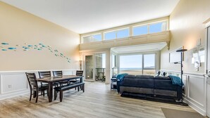 Un vaste espace de vie comprenant une table et des chaises, un canapé et une grande fenêtre donnant sur la plage.
