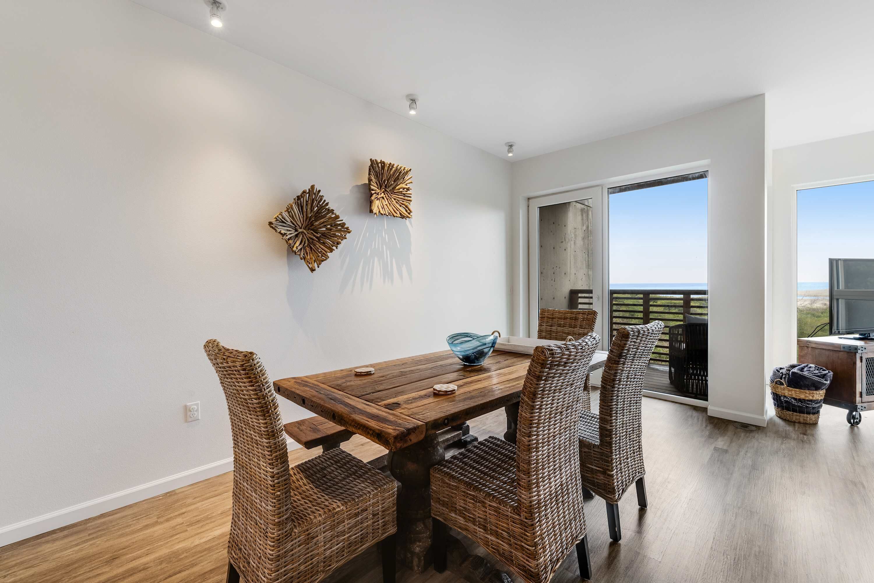 A dining area with a wooden table and wicker chairs, a blue glass bowl, and two wall art pieces.