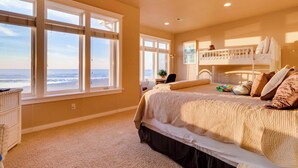 Een slaapkamer met een groot raam met uitzicht op het strand, een stapelbed en een bureau met een stoel.