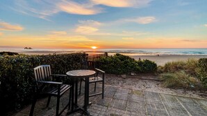 Een terras met twee stoelen en een tafel, met uitzicht op een strand bij zonsondergang.