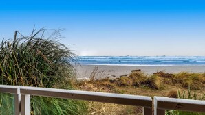 Vista a la playa con barandilla de madera, olas del océano y dunas cubiertas de hierba.