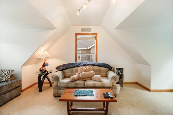 A cozy loft bedroom with a sloped ceiling, a large window, a sofa, a coffee table, and a bookshelf. - Comfortable home with a private game room blocks from the beach (Rockaway Beach)