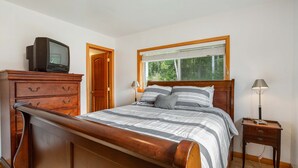 Una camera da letto con un letto di legno, una cassettiera, un televisore sopra e un comodino con una lampada. Si apre una finestra sulla vista degli alberi.