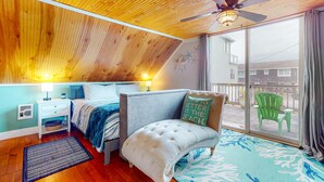 A bedroom with a sloped ceiling, a bed with a tufted headboard, a nightstand, a window with a view of the outside, and a ceiling fan. - Dog Friendly Cottage with Private Hot Tub Close to Beach and Downtown (Rockaway Beach)