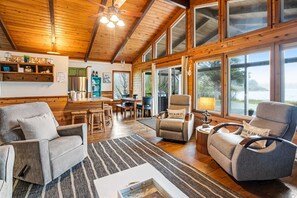 A spacious living room with wooden floors, a large window, and a view of the outdoors. - Rustic waterfront woodland lodge with impressive lake views from patio (Rockaway Beach)
