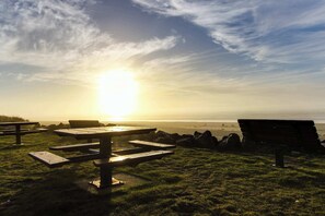 A picnic area with benches and picnic tables, overlooking a beach with the sun setting in the background. - Charming Cottage with Central Location Mountain Views and Beach Access Across Street (Rockaway Beach)