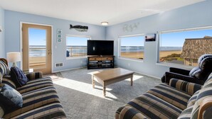 Een woonkamer met een televisie, bank en grote ramen met uitzicht op het strand.