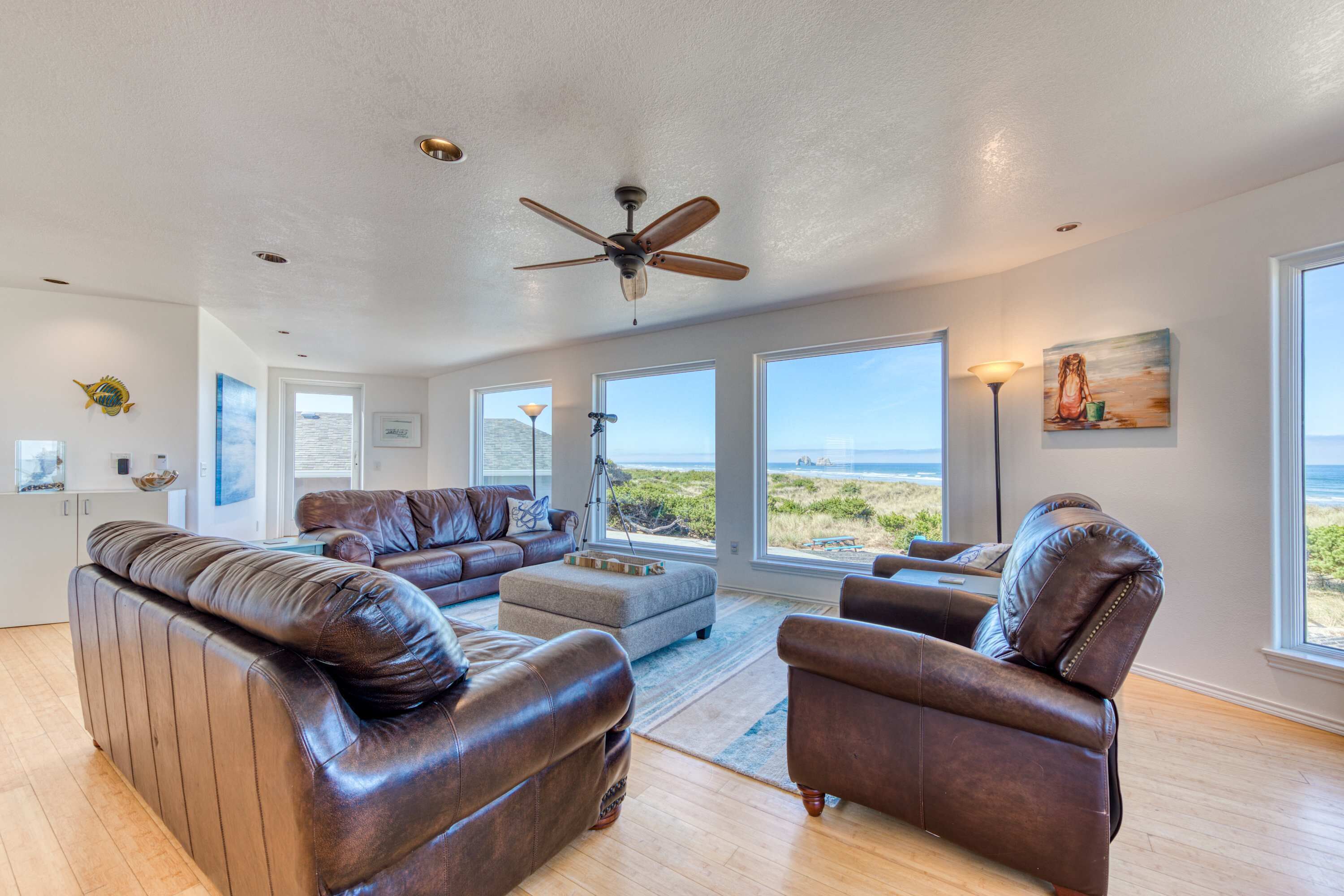 A spacious living room with leather sofas, a ceiling fan, large windows with a view of the ocean, and a painting on the wall.