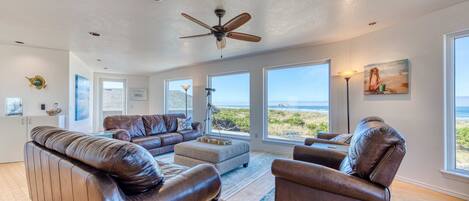 A spacious living room with leather sofas, a ceiling fan, large windows with a view of the ocean, and a painting on the wall.